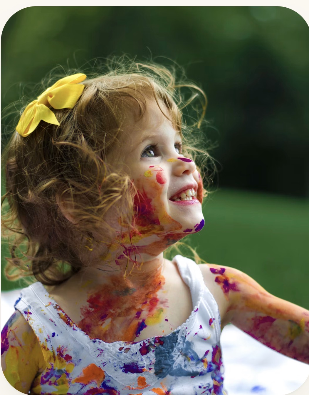 A joyful little girl with a yellow bow in her hair, smiling with colorful face paint, enjoying a creative outdoor activity